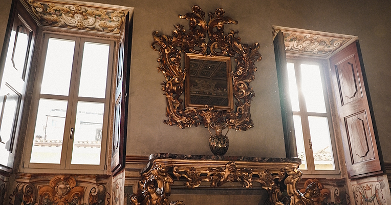 Baroque interior with gilded mirror, carved console and decorative frescoes between two bright windows.