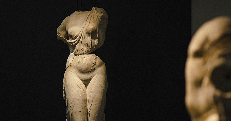 Classical female sculpture without head and arms, displayed in a museum against a dark background.