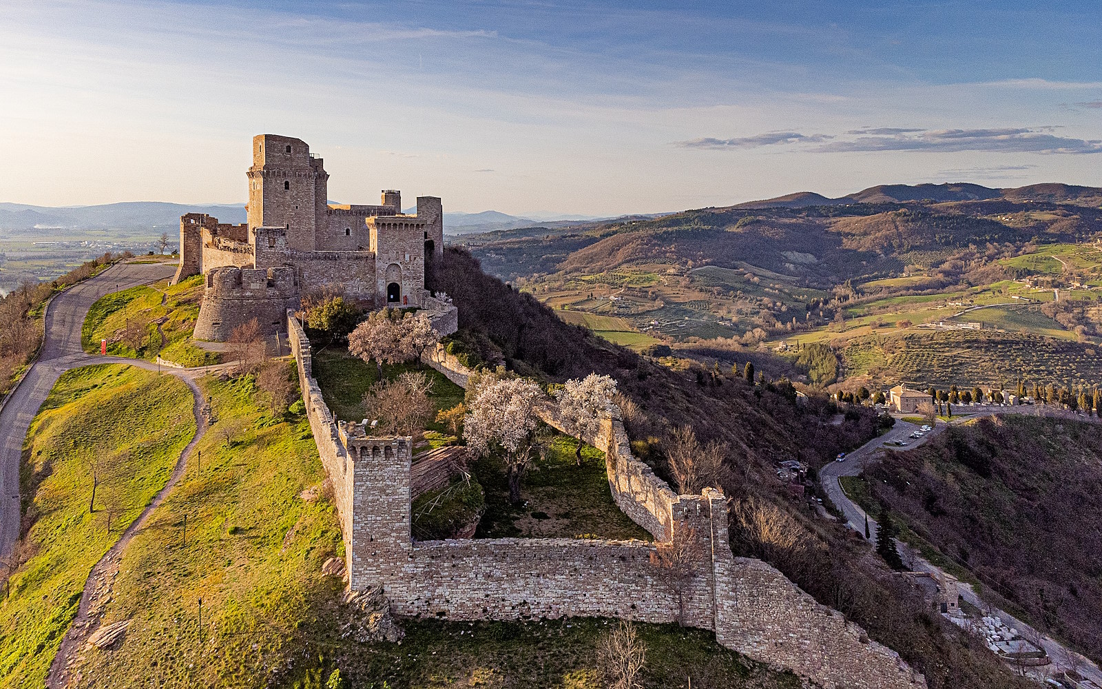 Aerial view of the Rocca Maggiore in Assisi, surrounded by medieval walls, overlooking the valley amid Umbrian hills.