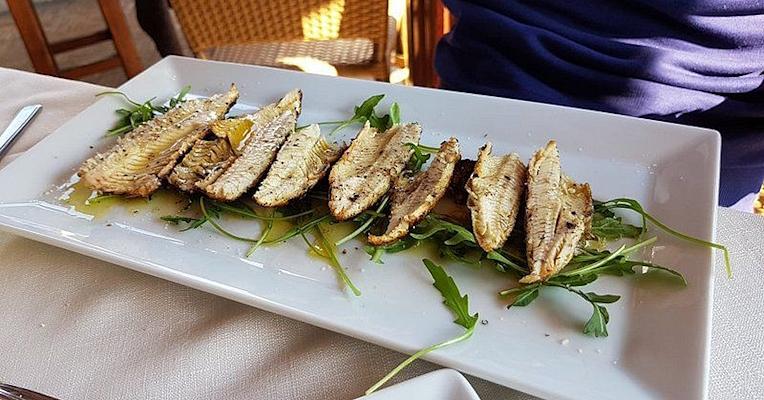 Filetti di pesce di lago grigliati (carbonaretti), serviti su letto di rucola con olio extravergine, impiattati su vassoio bianco.
