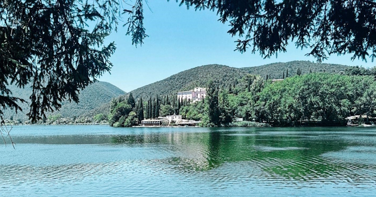  View of Lake Piediluco, with a staircase in the foreground on the left and a small boat moored on the shore to the right; in the background, wooded hills and a pink building nestled among cypress trees. 