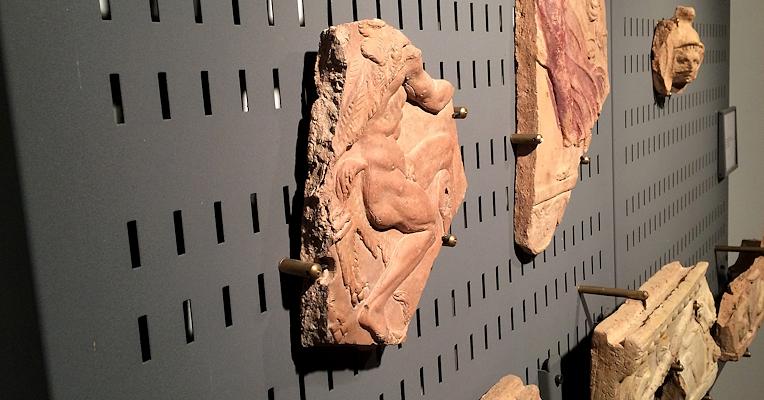 Fragments of decorated terracotta with human figures, displayed on a black perforated wall in an archaeological museum