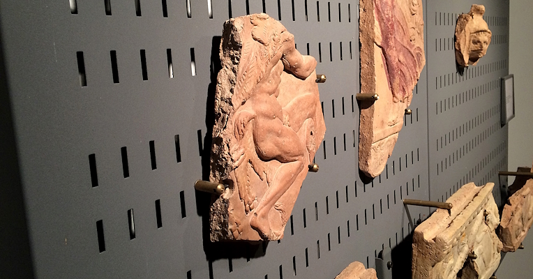 Frammenti di terracotta decorata con figure umane, esposti su una parete forata nera in un museo archeologico