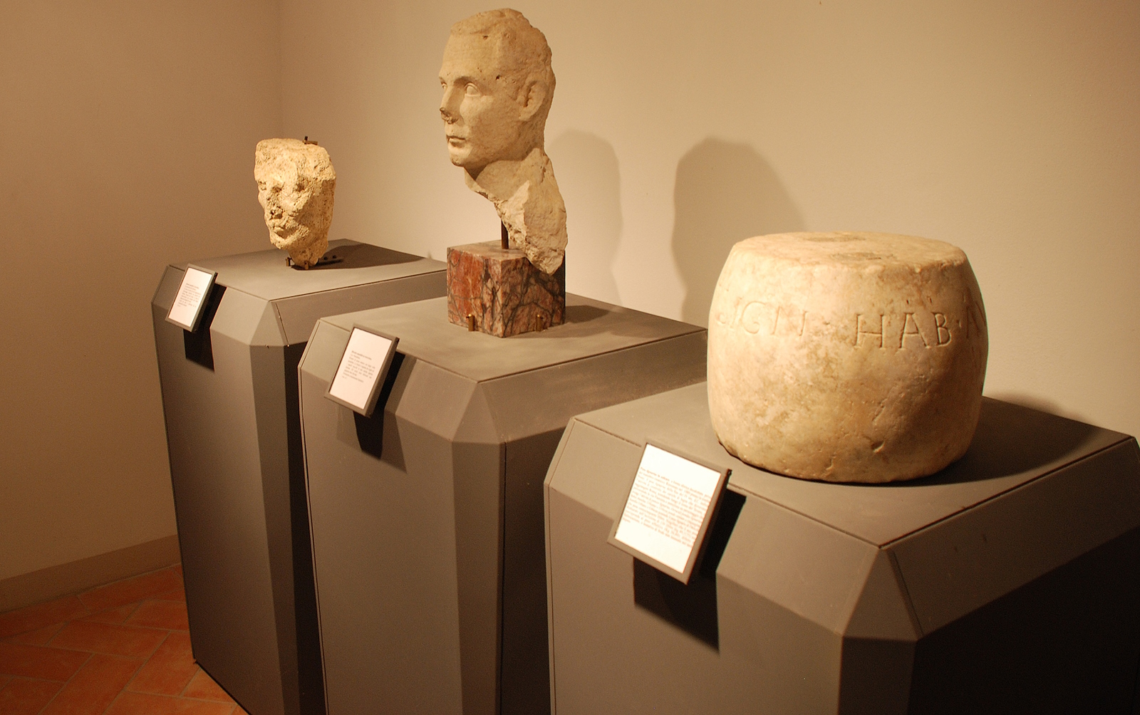 Two stone busts and a cylindrical object with Latin inscription, displayed on pedestals with captions