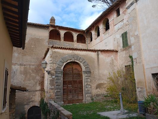 Ancient stone building with a large arched portal and upper loggia, surrounded by plastered walls and a grassy garden