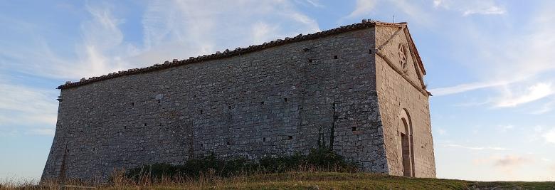 Immagine: Side view of a stone building located on a grassy hill, with a blue sky and light clouds in the background. 