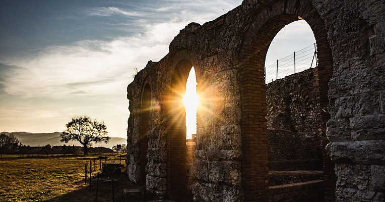 Sonne, die hinter einem Bogen des römischen Theaters von Gubbio untergeht und warme Licht- und Schattenspiele über die umgebende Landschaft wirft.