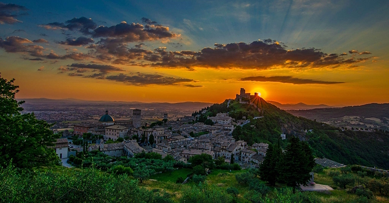  Panoramic view of Assisi at sunset, with the sun setting behind a hill and the sky tinged with orange and blue. 