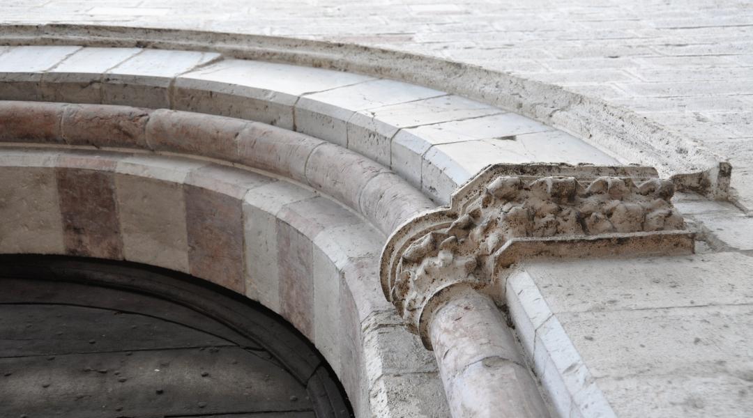 Detail eines romanischen Steinbogens der Kirche San Biagio mit zweifarbigen Steinbändern in abwechselnden Streifen. Der Bogen wird von einer Säule mit einem Kapitell getragen, das mit floralen Skulpturen verziert ist.
