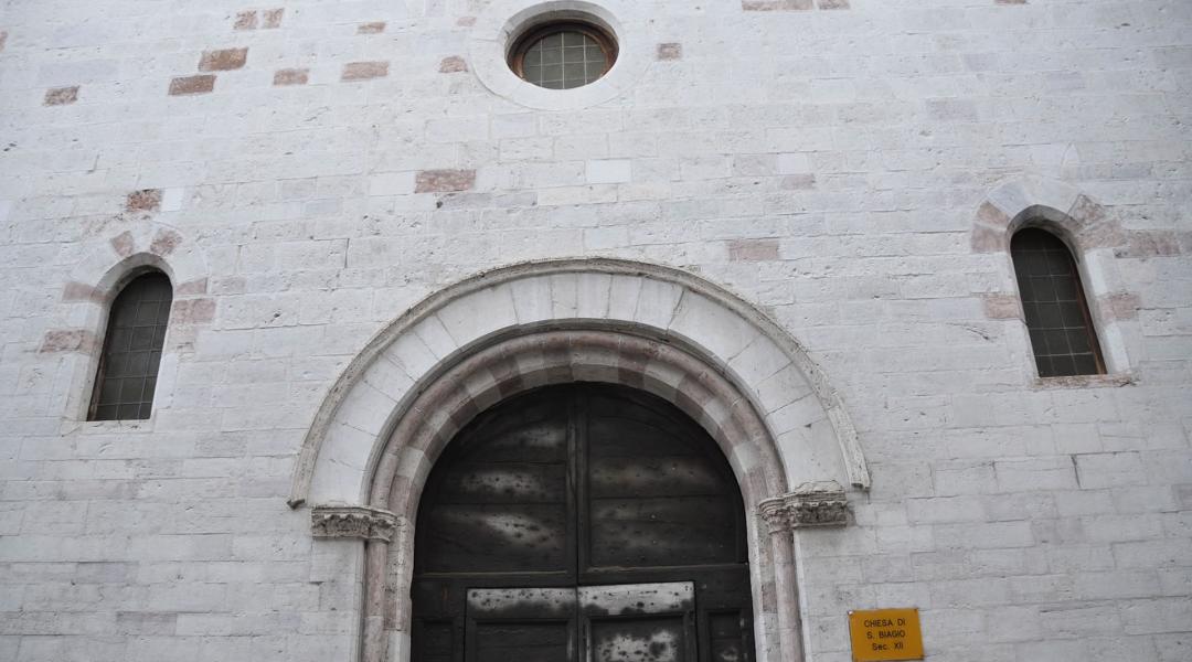 Fassade der romanischen Kirche San Biagio aus weißem und rosafarbenem Stein, mit einem zentralen Rundbogenportal. Zwei schmale Rundbogenfenster flankieren das Portal, und ein kleines rundes Fenster ist oben angebracht.
