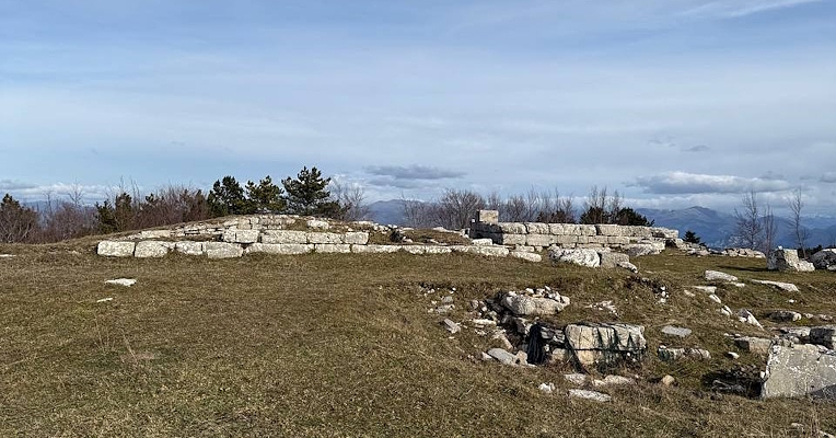 Stone ruins on a grassy hill with a panoramic view of hills and mountains on the horizon under a cloudy sky.