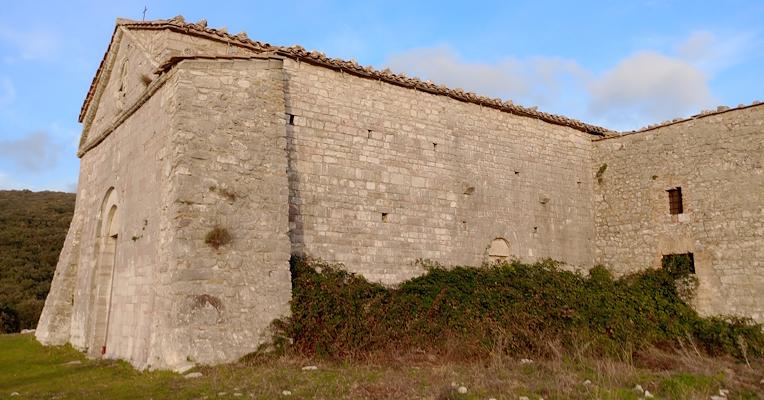 Ancien bâtiment en pierre, entouré de végétation et de collines, sous un ciel bleu avec quelques nuages épars.