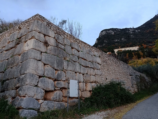 Ancient stone block wall along a mountain road, with vegetation and hills in the background.