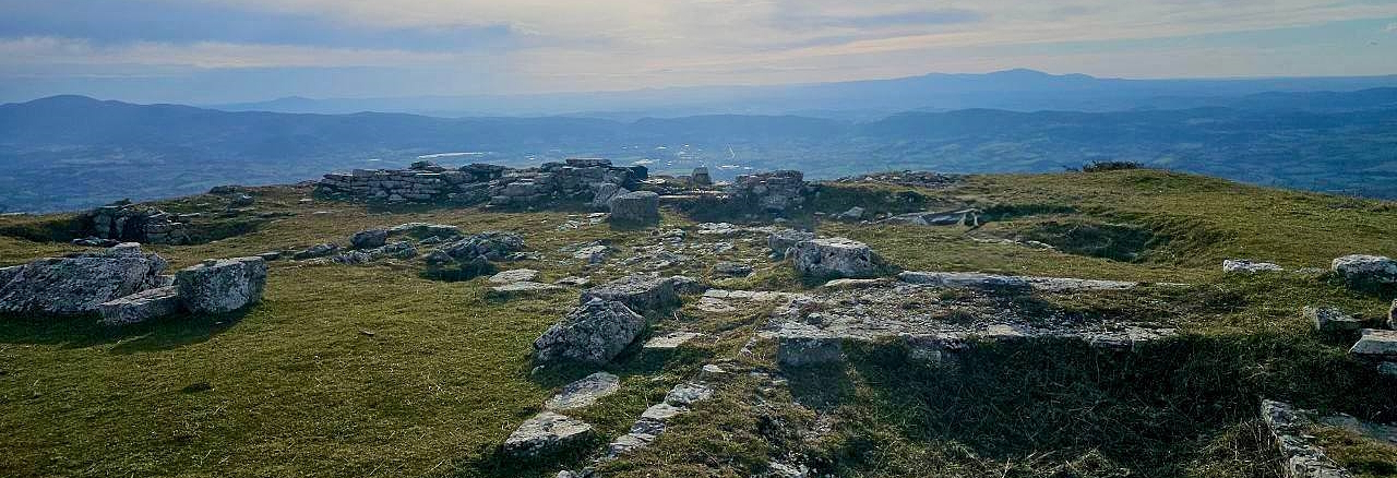 Stone ruins on a grassy hilltop with a panoramic view of hills and mountains on the horizon under a cloudy sky.