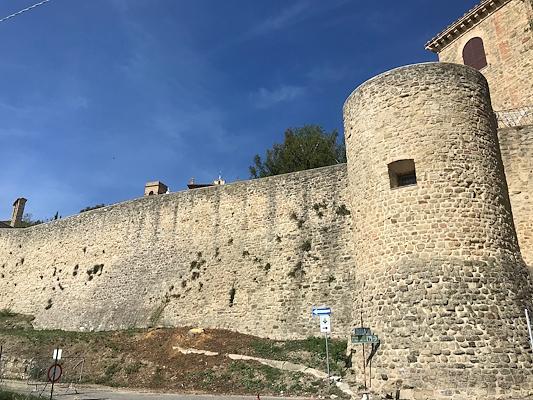 Ancient stone walls with a cylindrical tower in Gualdo Cattaneo, photographed from below under a clear sky.