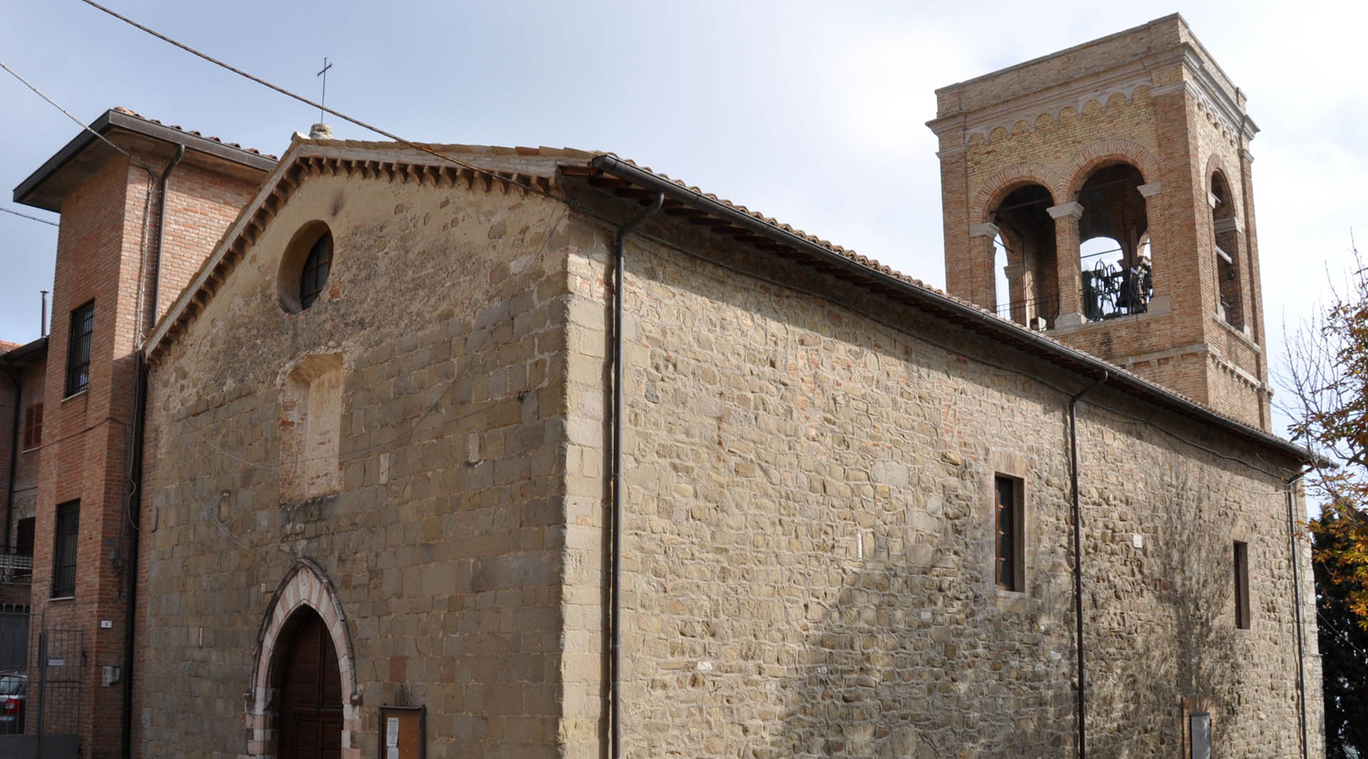 Stone church with a simple façade and a brick bell tower, seen from the side under a cloudy sky.