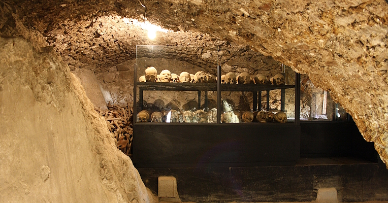 Unterirdische Krypta mit menschlichen Schädeln und Knochen in Glasvitrinen, beleuchtet von einer einzelnen Glühbirne.