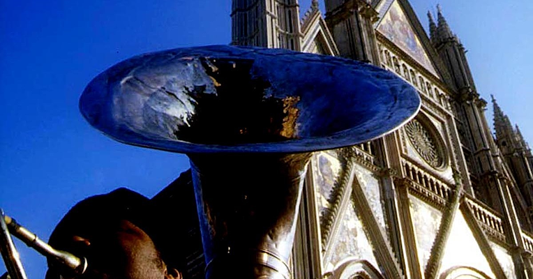 Homme jouant du tuba, vu d’en bas, avec la façade de la cathédrale d’Orvieto et un ciel bleu clair en arrière-plan.