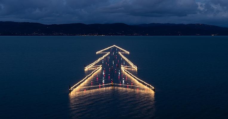 Albero di Natale illuminato galleggiante sul Lago Trasimeno al tramonto, riflesso nelle acque tranquille.