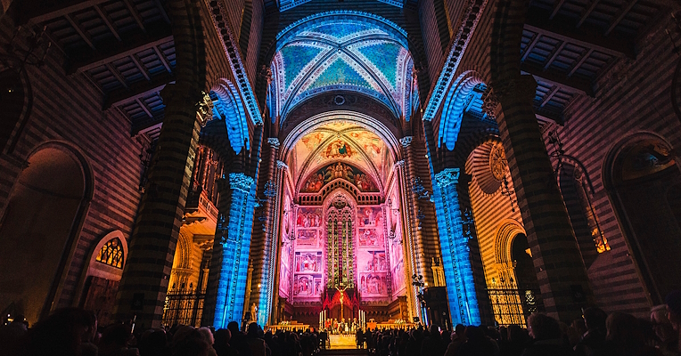 Interior of Orvieto Cathedral lit by colorful lights during a concert, with the audience seated along the aisles