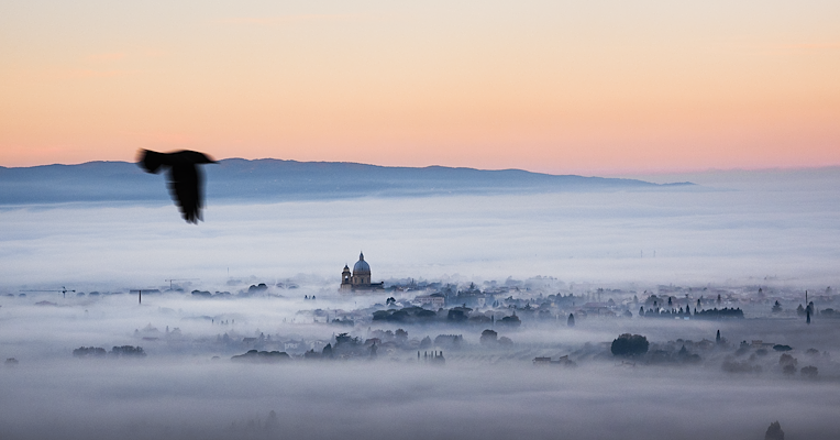 The Umbrian Valley wrapped in evening mist, with the dome of the Basilica of Santa Maria degli Angeli in Assisi standing out in the background.