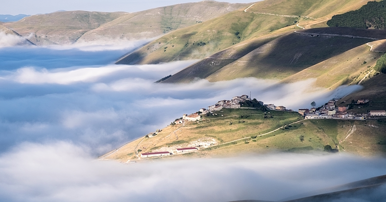  A village on a hill, surrounded by low-lying clouds amid green slopes and peaks bathed in the morning light. 