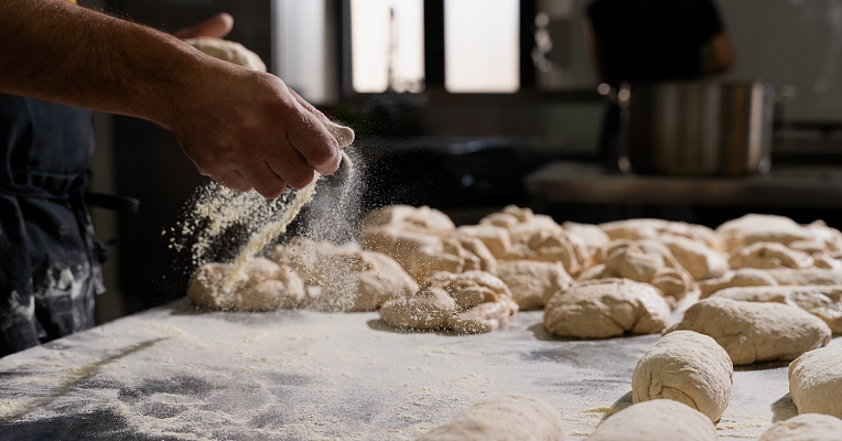 Unbaked bread rolls arranged on a wooden board, as a skilled hand dusts them with flour.