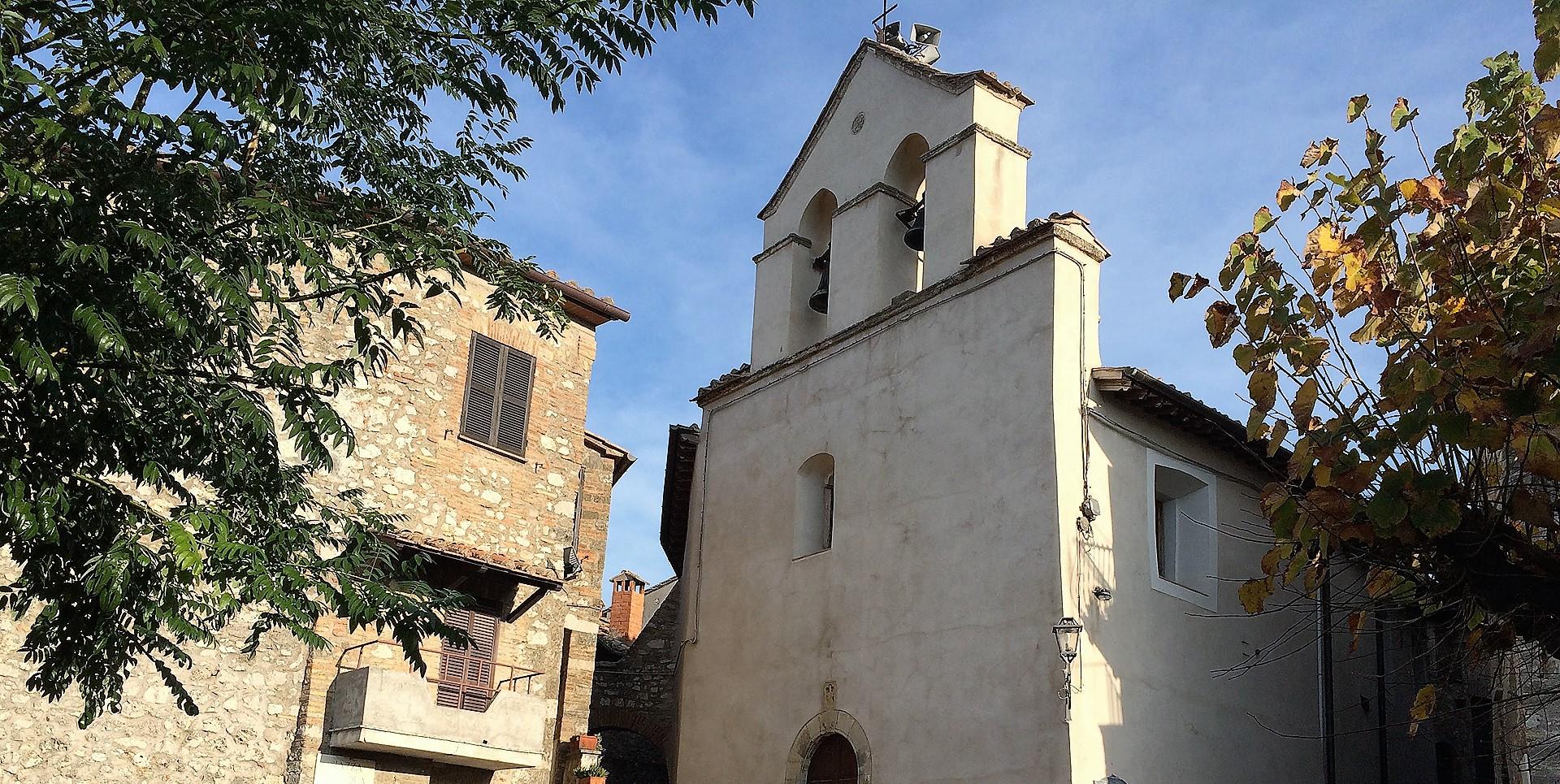 Church with a bell-gable and stone staircase, next to a stone house with shutters and a flowered balcony.