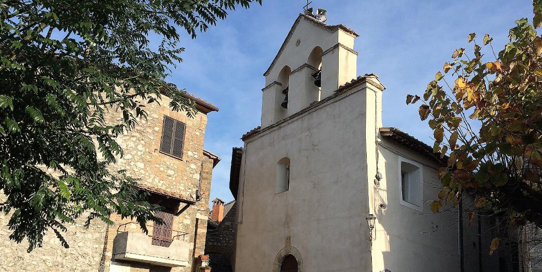 Church with a bell-gable and stone staircase, next to a stone house with shutters and a flowered balcony.
