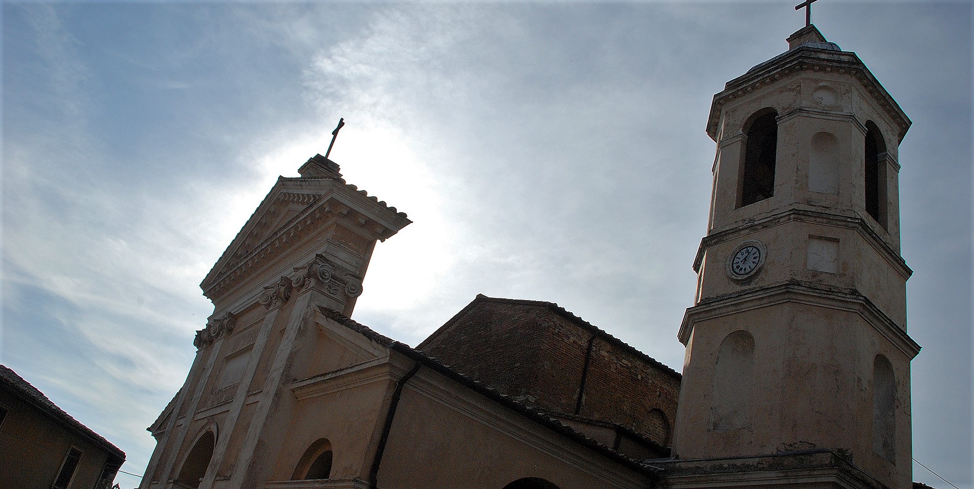 Façade and bell tower of the Collegiata of Santa Maria Assunta in Otricoli, backlit against a hazy sky.
