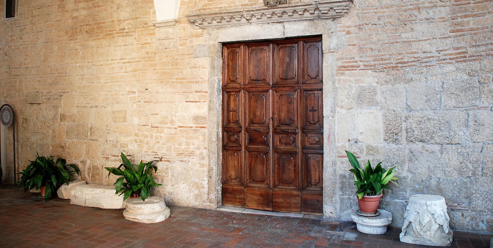 Wooden portal of the Collegiata, framed by stone walls and flanked by potted plants.