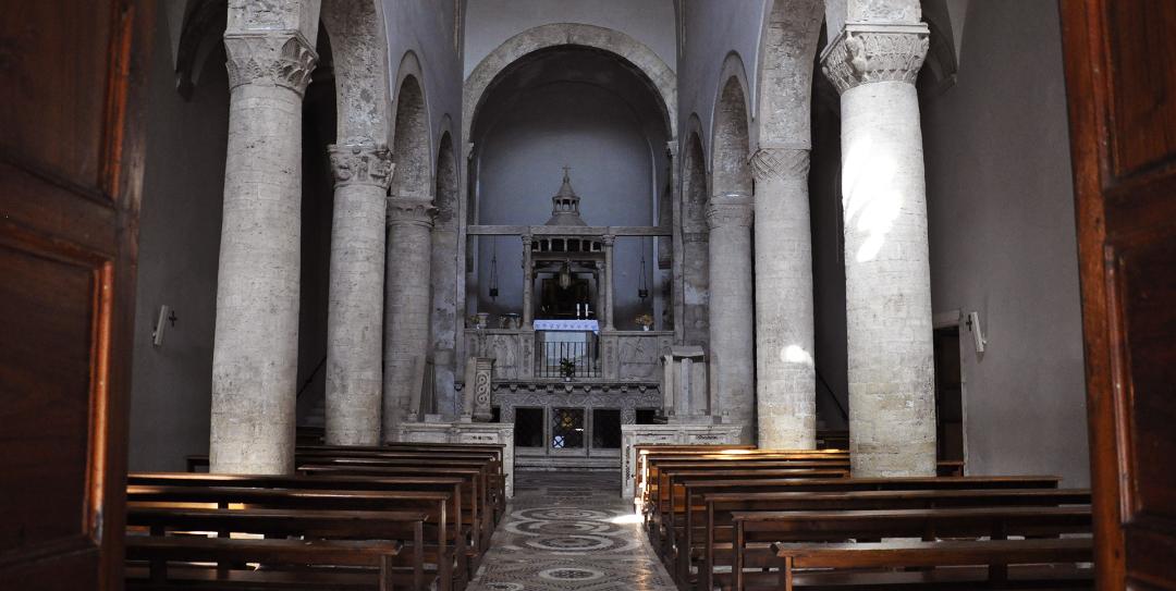 Interno e navata centrale della chiesa di Santa Maria Assunta con colonne marmoree, banchi in legno e altare rialzato in fondo.