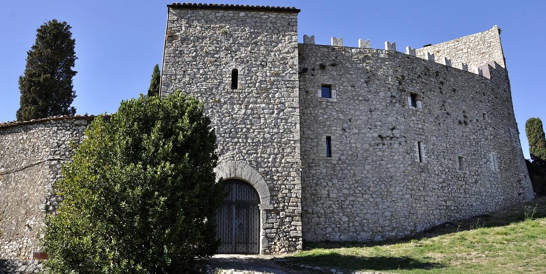 Façade en pierre du Castello del Poggio avec tour crénelée, portail en arc et remparts fortifiés, entourée de verdure.