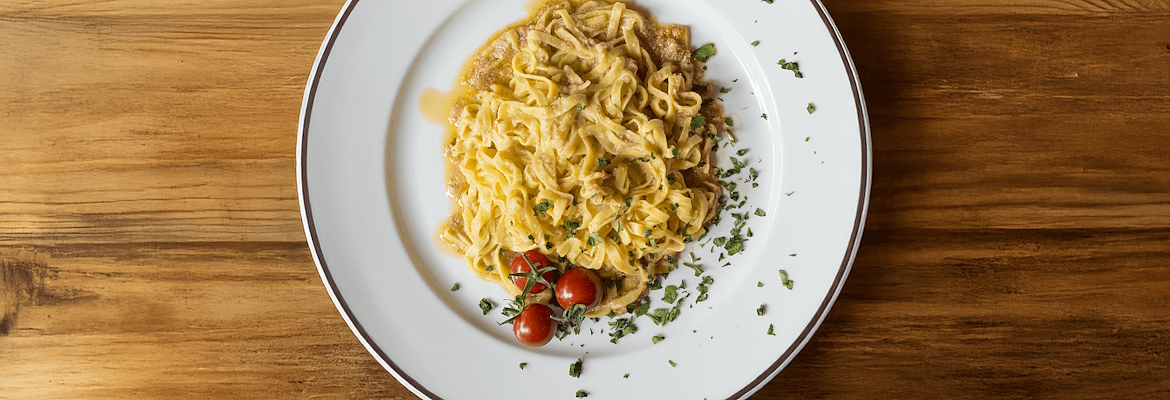 Tagliatelle with smoked tench, served with aromatic herbs and cherry tomatoes on a white plate, placed on a wooden table.
