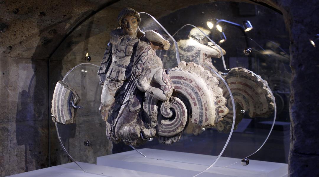 Remains of a polychrome terracotta sculpture depicting a human figure with intricate decorative details. The sculpture is exhibited in an illuminated case, highlighting the vibrant colors and complex decorations of the artifact.