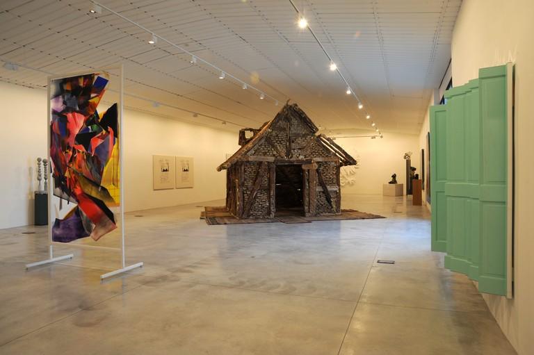Una sala espositiva con una scultura di una capanna al centro, un’opera astratta a sinistra e una finestra verde a destra.
