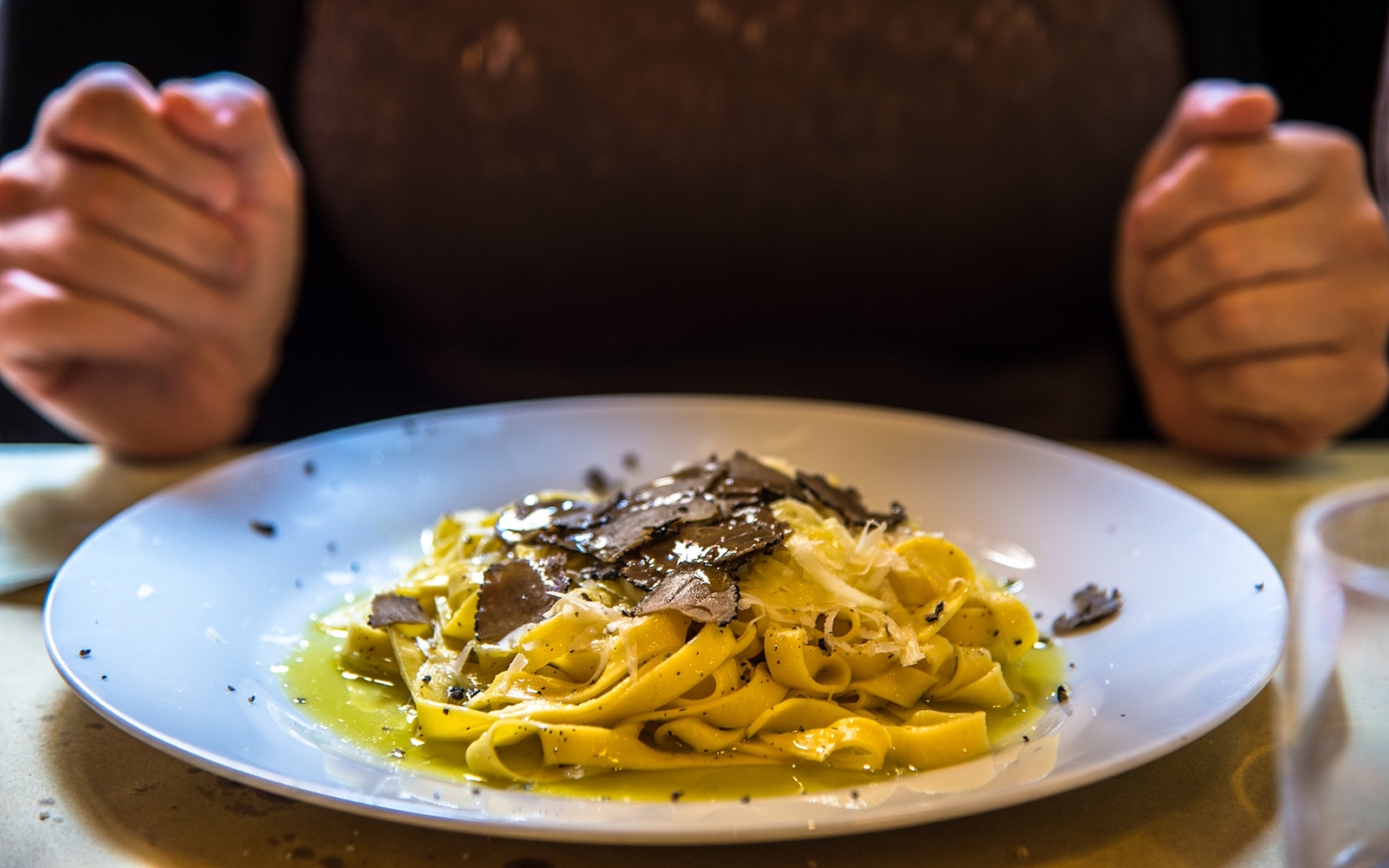 Plate of tagliatelle with truffle shavings, with hands resting on the table ready to eat in the background.