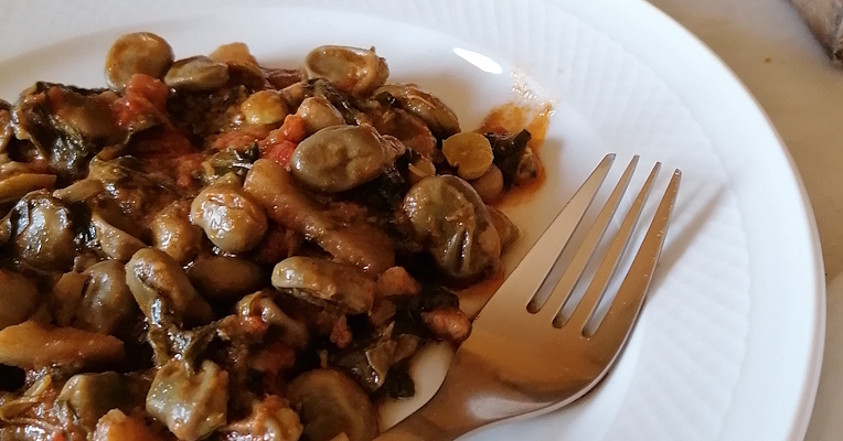 Plate of stewed fava beans with tomato and herbs on a white plate, with a fork in the foreground and bread in the background.
