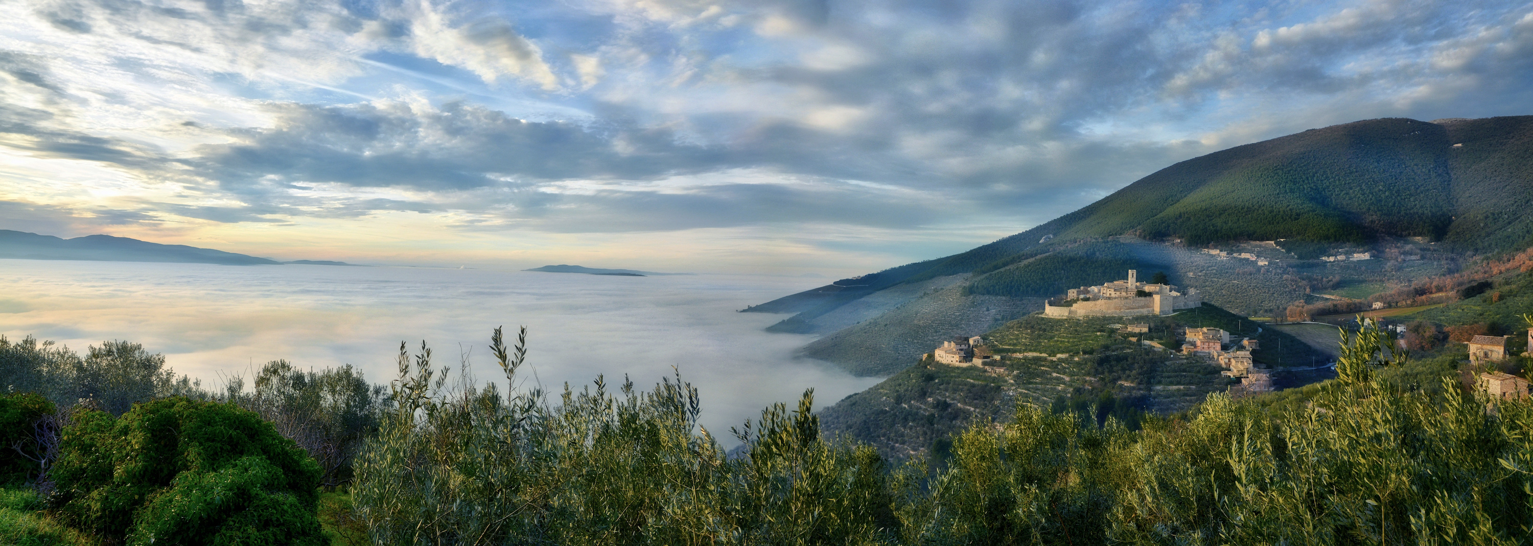 Panoramic view of the Umbrian hills with the Castle of Campello Alto surrounded by olive groves and wrapped in a blanket of mist.