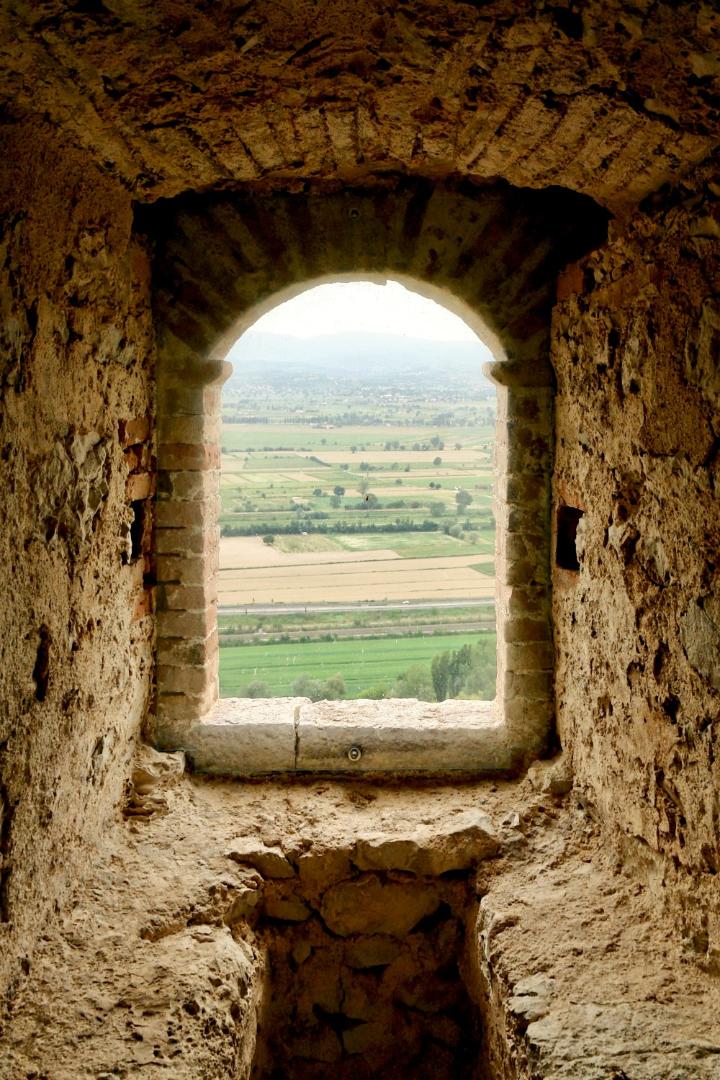 Interno in pietra del castello con una stretta finestra ad arco che incornicia la vista della Valle Umbra, tra campi coltivati e colline lontane