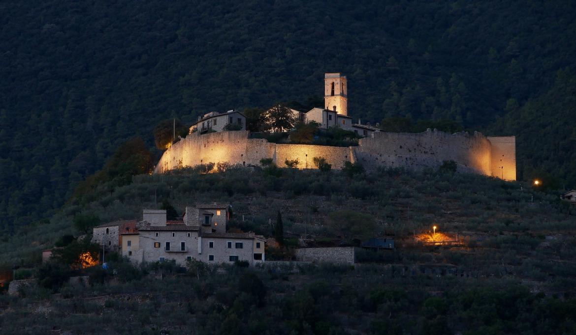 The fortified village of Campello Alto illuminated at dusk, with its walls and bell tower rising above a hillside covered in olive trees