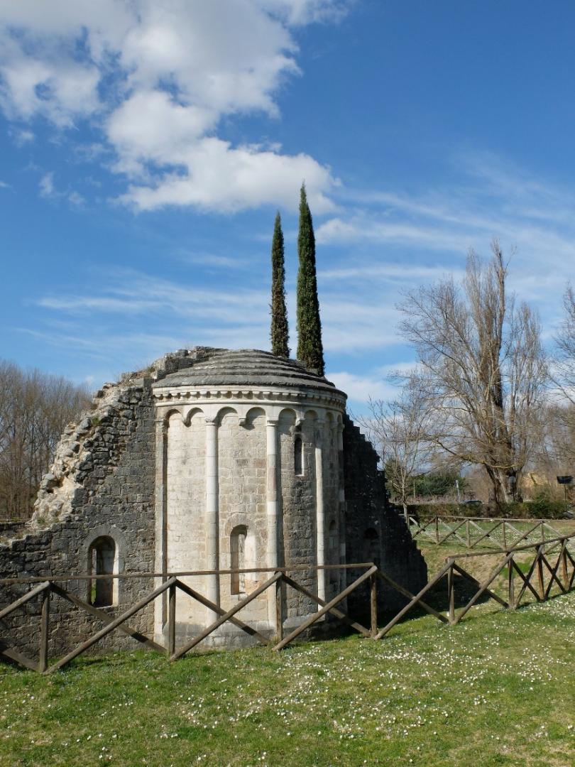 Romanesque stone apse rising from a green meadow, with two cypress trees in the background under a blue sky.