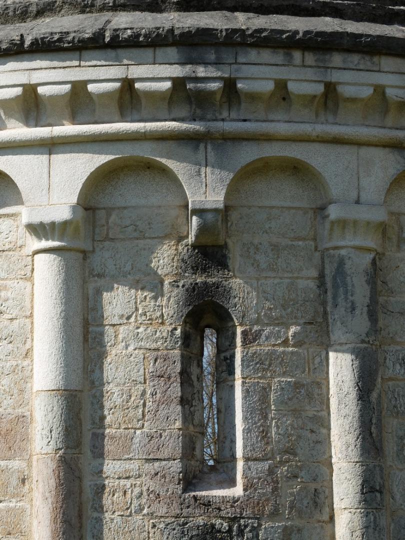 Close-up of the Romanesque apse with small arches and a narrow slit window, its pale stones lit by the sun