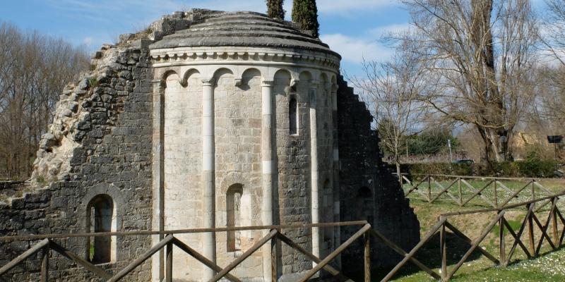 Abside romanica in pietra che emerge da un prato verde, con due cipressi sullo sfondo sotto un cielo azzurro.