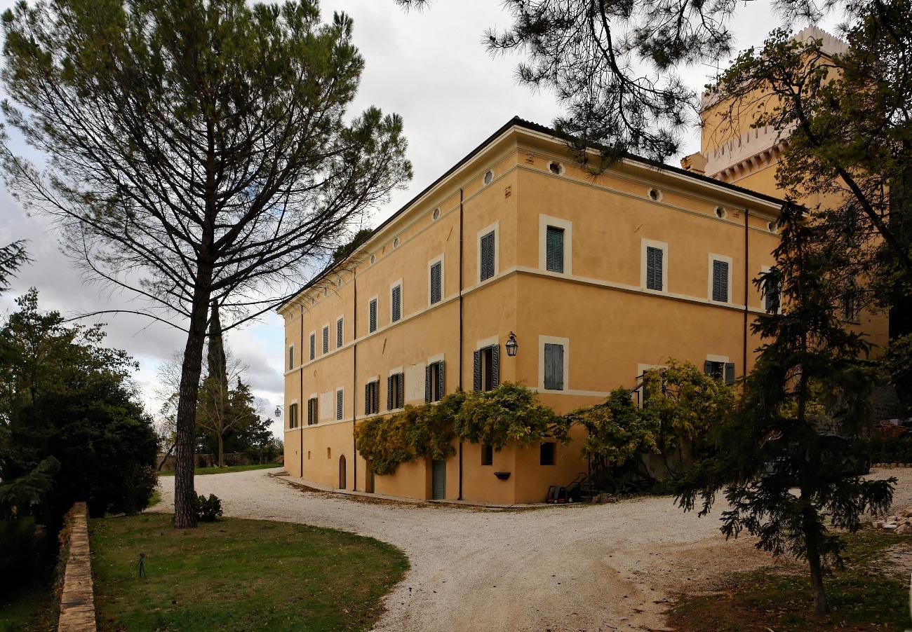 Large historic ochre-colored villa with tower and gravel driveway, surrounded by trees and gardens