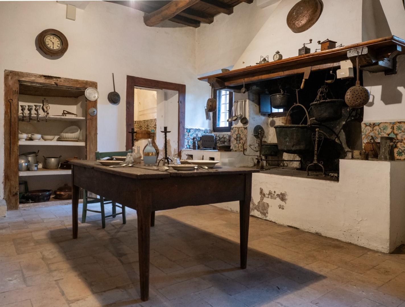 Rural kitchen with hearth, iron pots, shelves and a table set with dishes.