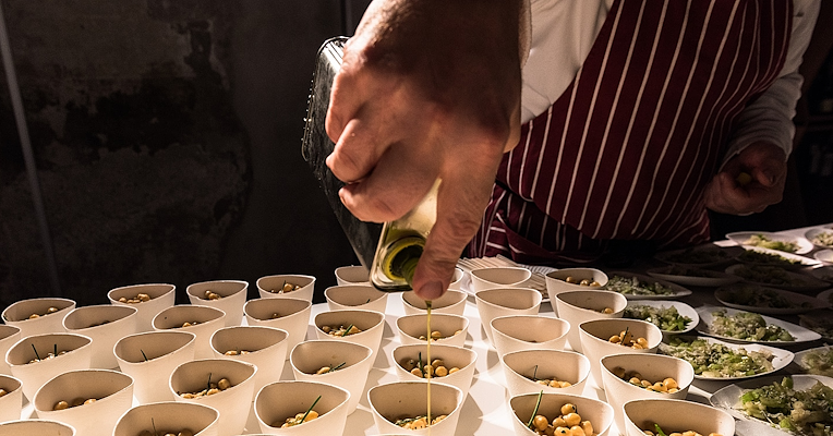 A chef pours extra virgin olive oil over small portions of legumes served in cups during a food tasting.
