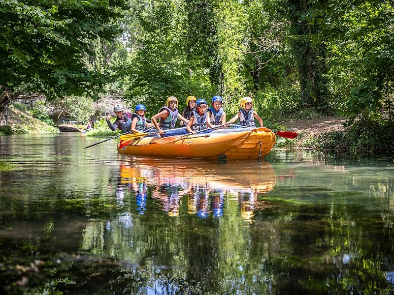 Group of people rafting on an orange inflatable boat along a river surrounded by vegetation, reflected on the water